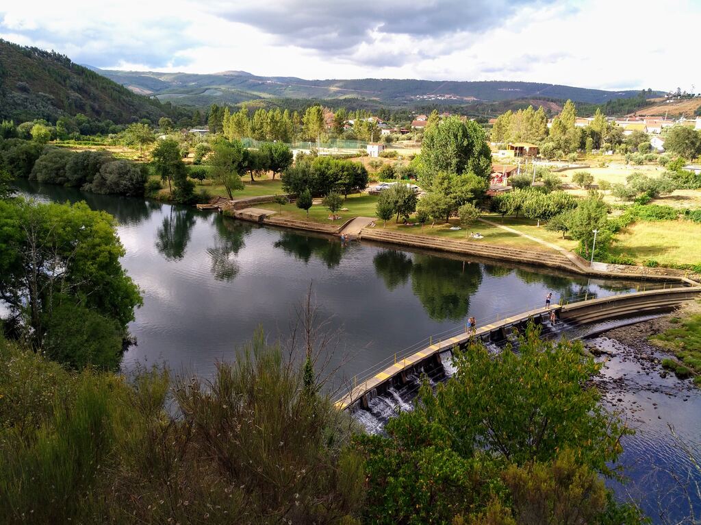 O rio Zêzere junto a Janeiro de Cima, visto do alto: o açude, o relvado do parque fluvial e a aldeia ao fundo, entre serras.
