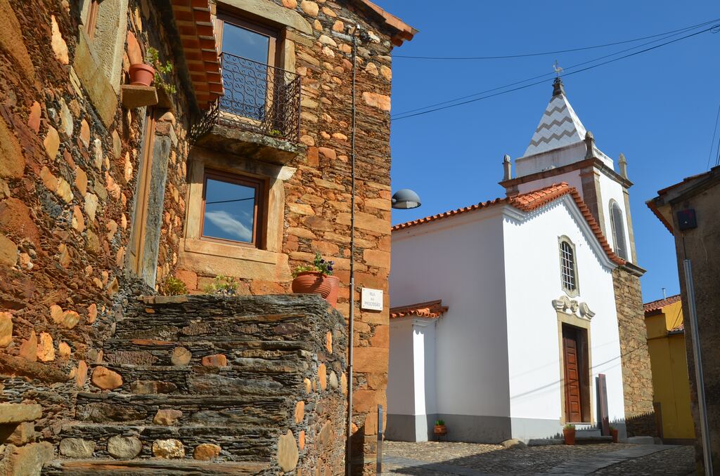 Casa de xisto com escada de lajes e varanda de ferro, ao lado de uma igreja branca com torre em bico, no centro de Janeiro de Cima.